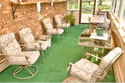 A sunroom with green carpet flooring and brick walls, furnished with six floral-patterned cushioned swivel chairs arranged around two glass-top tables. The room has large windows allowing natural light and a view of the outdoor greenery.