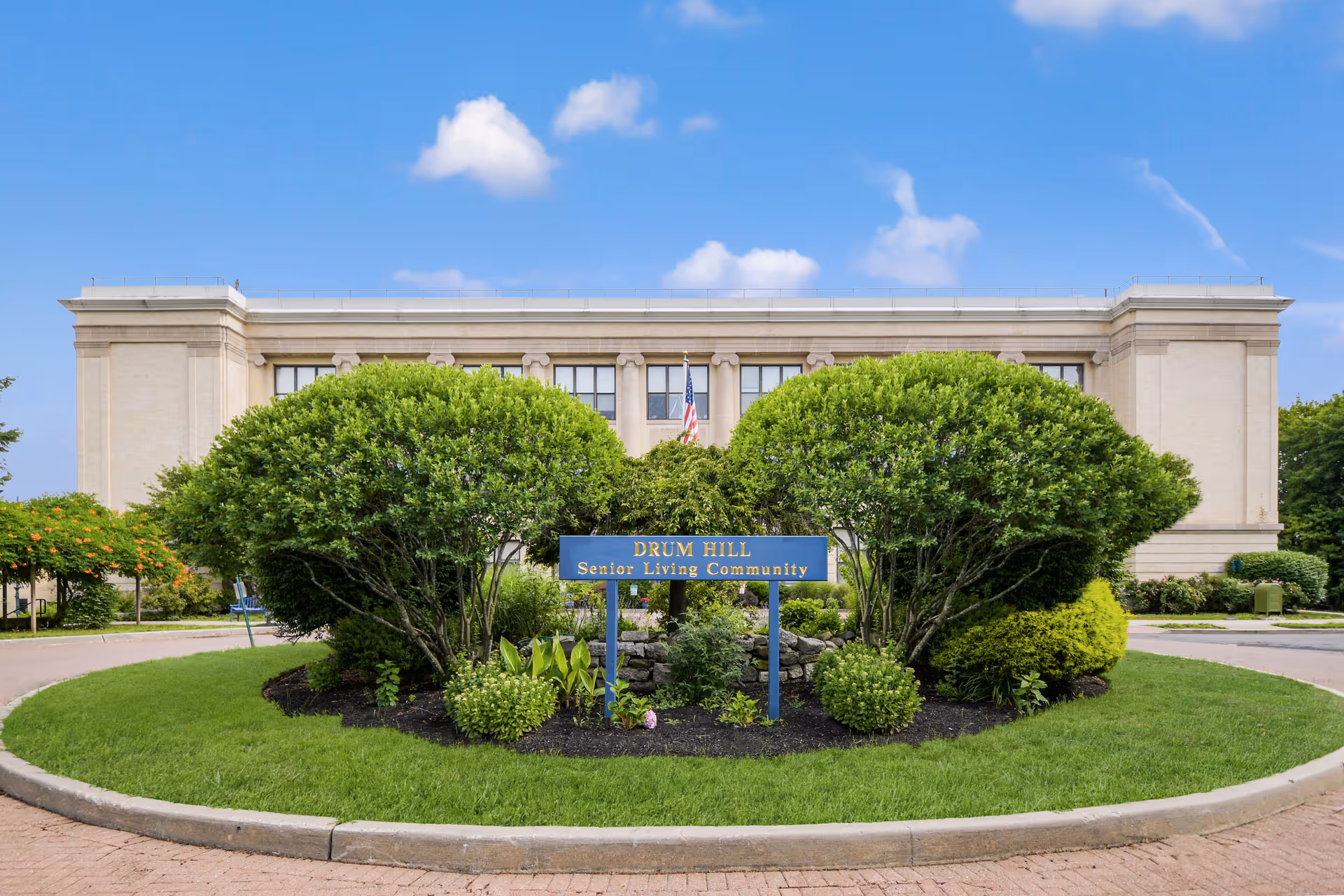 Front view of the Drum Hill Senior Living Community building with trimmed shrubs and a blue sign centered in a circular driveway.