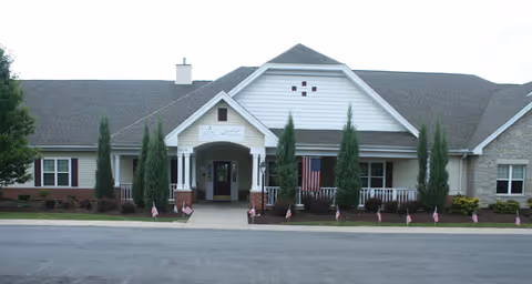 Front exterior view of a single-story senior living facility building with a covered entrance, white siding, brick accents, and several small American flags lining the walkway.