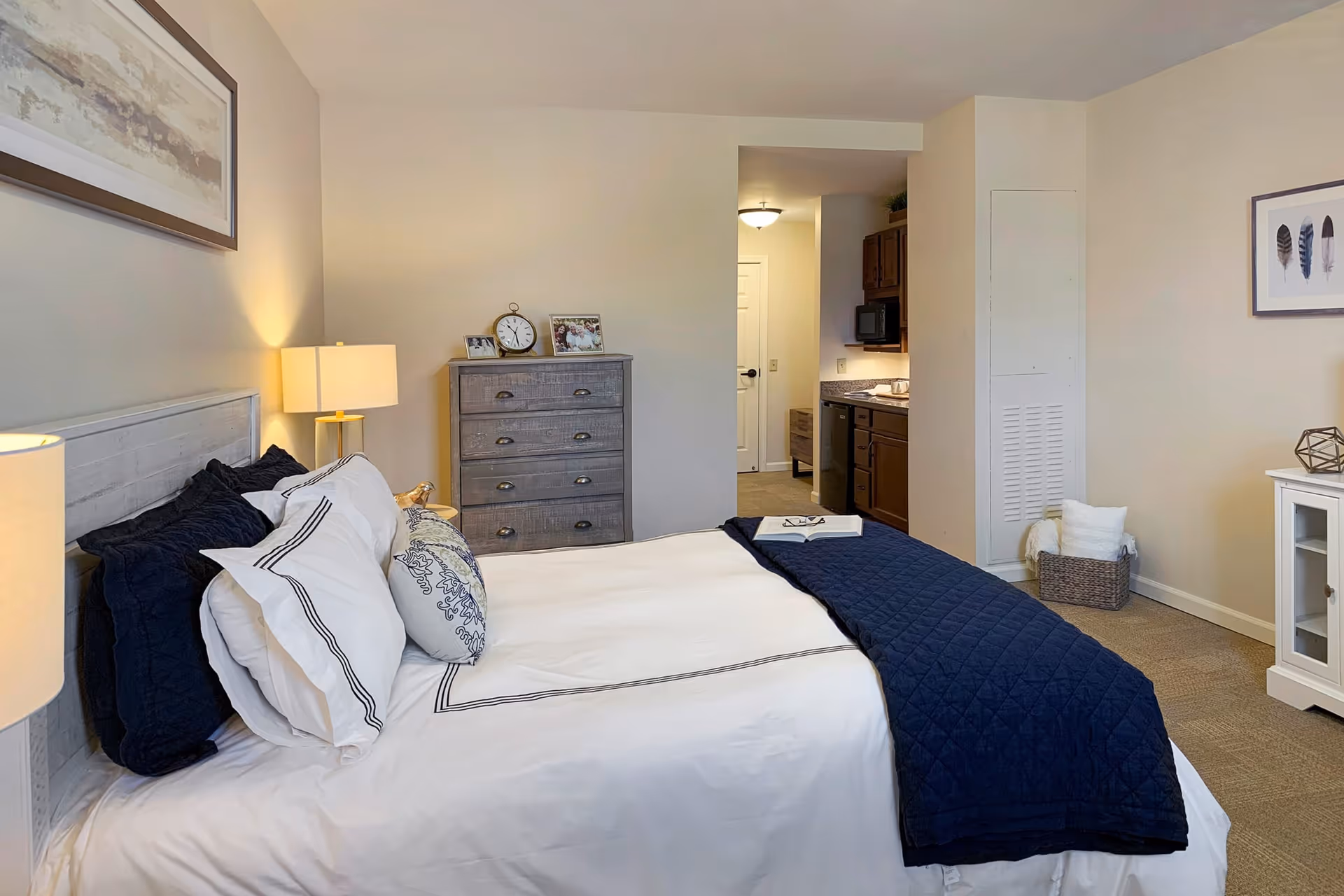A cozy bedroom in a senior living facility featuring a neatly made bed with white and navy blue bedding, a wooden headboard, and multiple pillows. To the left of the bed is a nightstand with a lamp. Across from the bed is a gray chest of drawers with a clock and framed photos on top. The room has beige walls and carpeted flooring. In the background, there is a small kitchenette area with dark wood cabinets and a microwave, and a hallway leading to a door. On the right side of the room, there is a white cabinet with decorative items and a basket with towels on the floor.