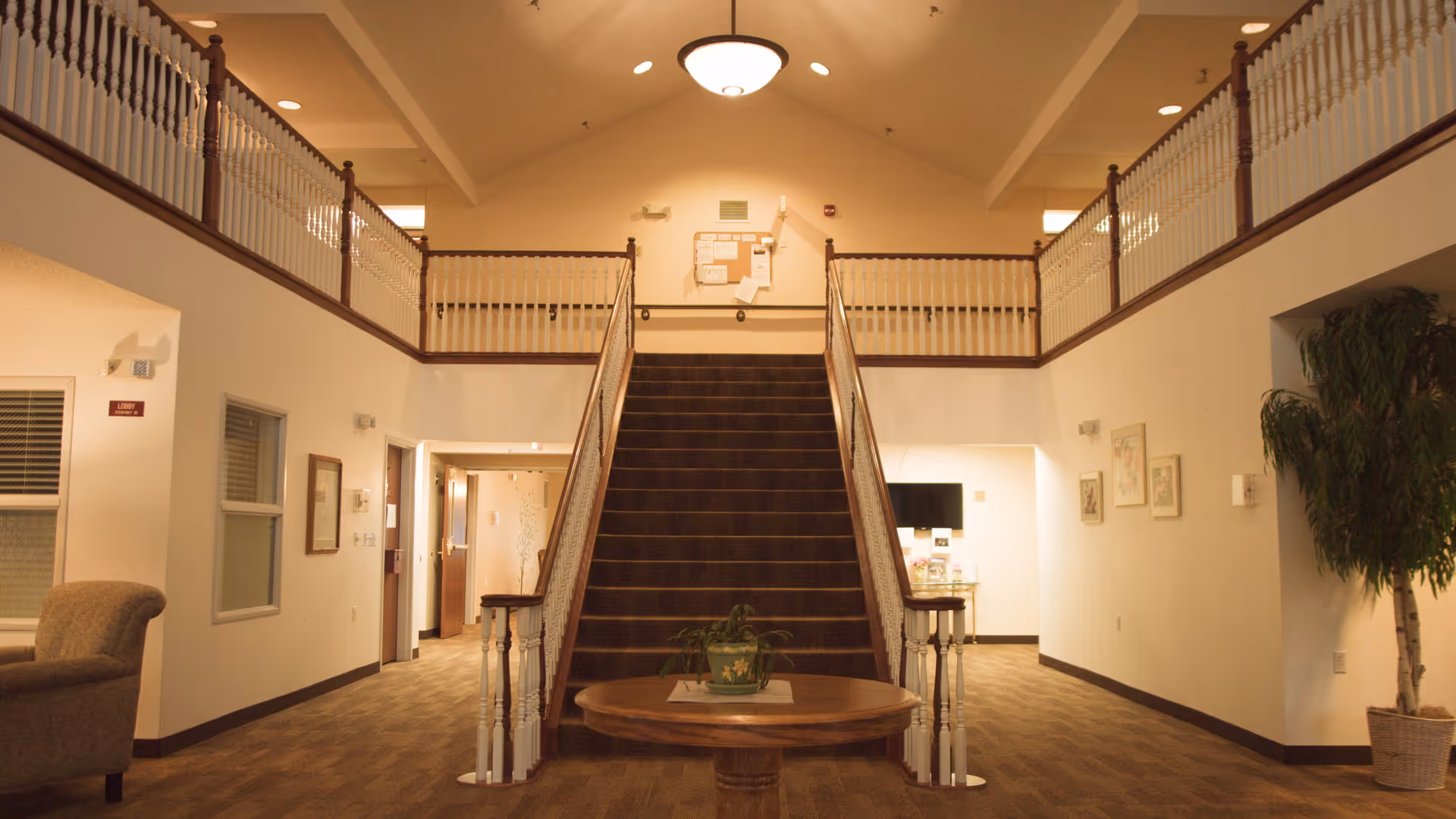 Interior view of a senior living facility lobby with a central staircase leading to an upper floor balcony. The space features beige walls, carpeted floors, a round wooden table with a potted plant in the center, framed artwork on the walls, and a large potted plant on the right side.