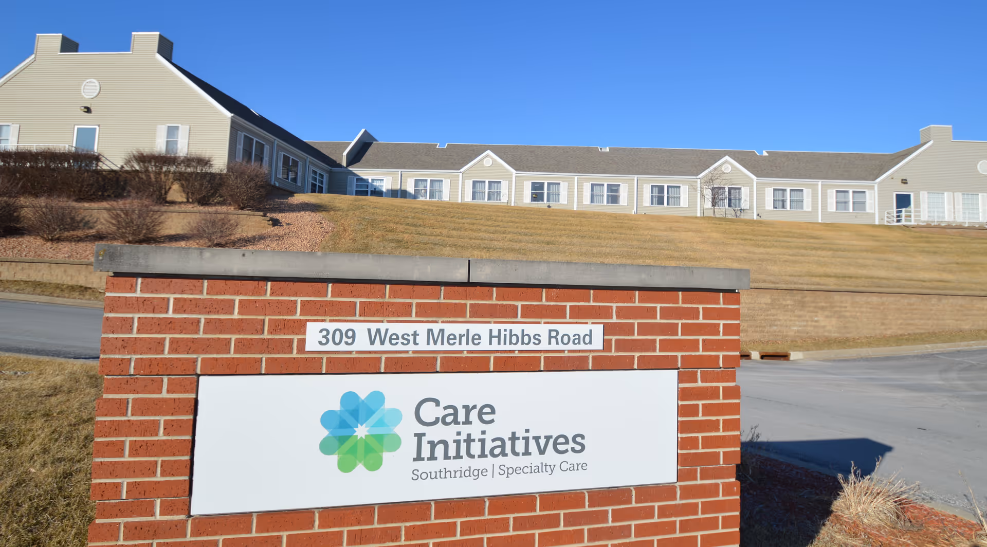 Exterior view of a senior living facility building on a grassy hill under a clear blue sky. In the foreground, there is a red brick sign with the address '309 West Merle Hibbs Road' and a white panel displaying the logo and name 'Care Initiatives Southridge Specialty Care.'