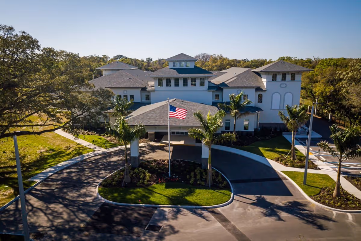 Front exterior view of a large, white senior living facility building with multiple roof peaks, surrounded by palm trees and greenery. A circular driveway with a flagpole flying the American flag is visible in front of the entrance.