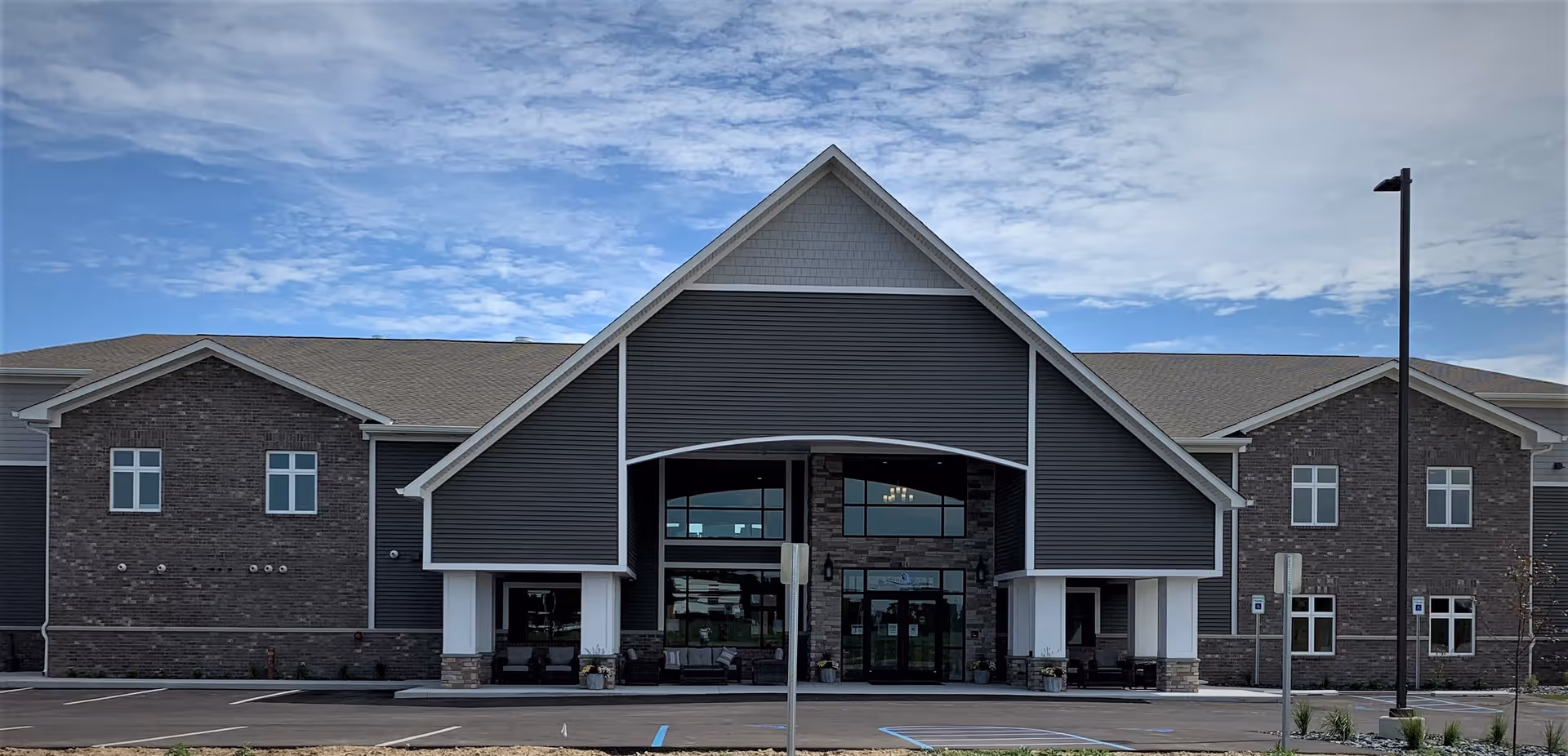 Front exterior of a two-story senior living building with a large peaked covered entrance, brick and gray siding, and a parking lot in front.