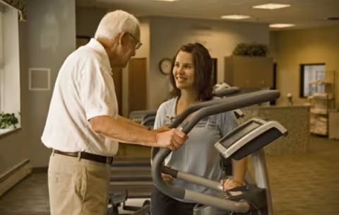 An elderly man using an elliptical machine in a fitness area while a female staff member or trainer stands beside him, smiling and providing assistance. The room has exercise equipment and a warm, well-lit interior.