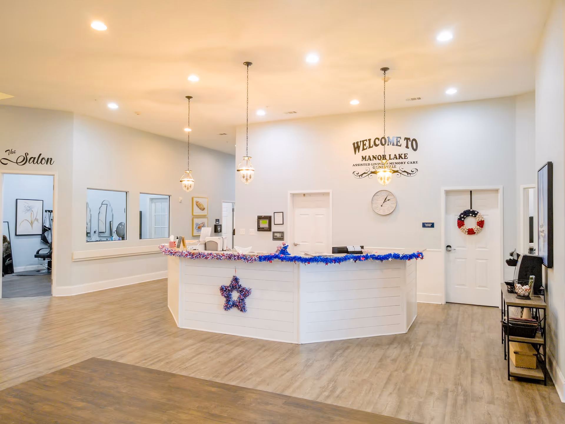 Reception area of Manor Lake Assisted Living & Memory Care - Gainesville with a white front desk decorated with red, white, and blue garlands and a star ornament. Behind the desk is a wall with a clock and a welcome sign. To the left is an entrance to The Salon, and to the right are white doors, one decorated with a patriotic wreath. The floor is wood-style, and the ceiling has recessed lighting and hanging pendant lights.