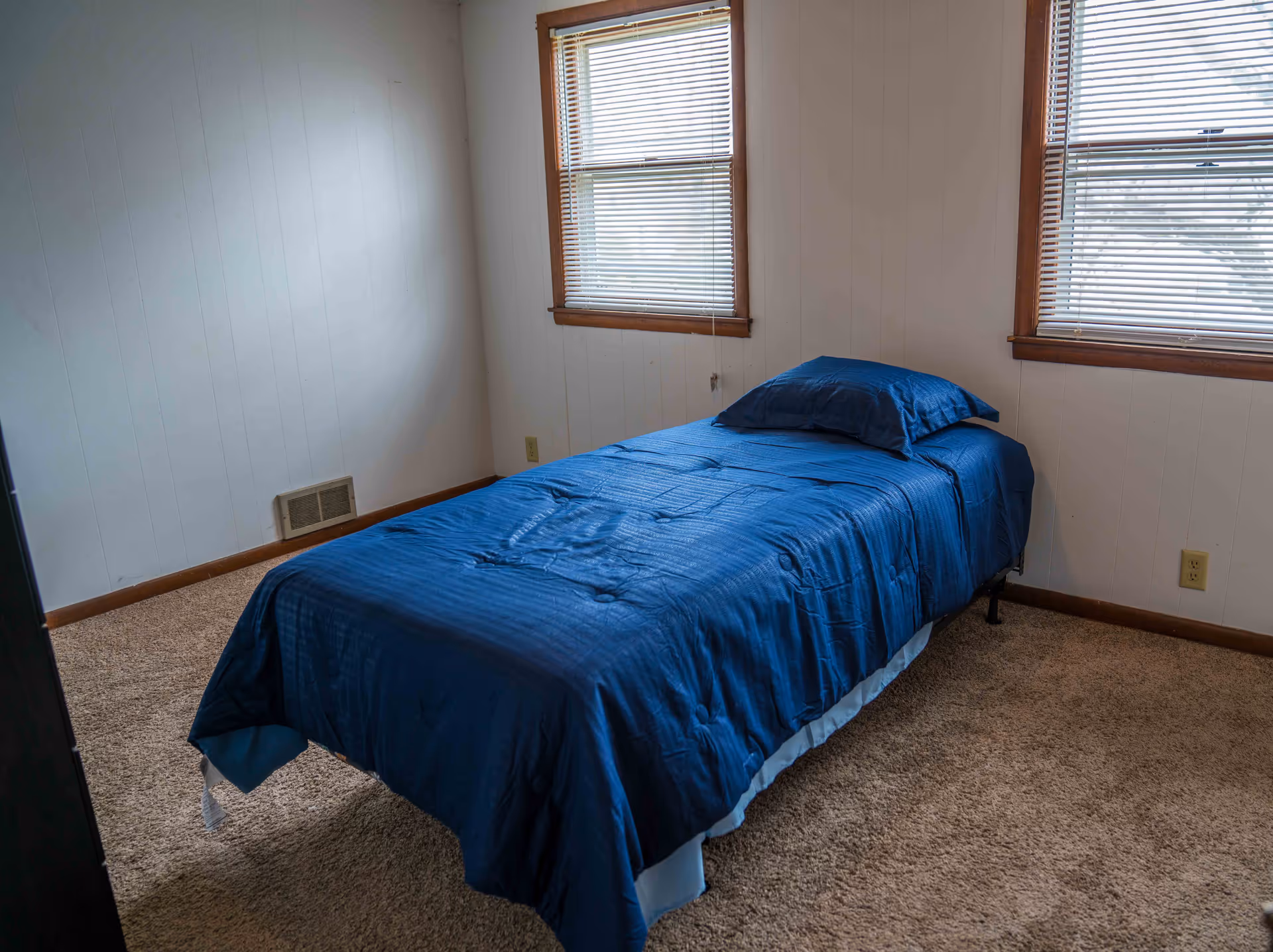 A simple carpeted bedroom with a single bed dressed in blue bedding beneath two windows with blinds.