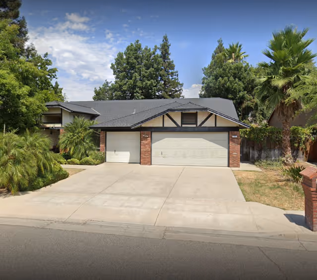 Front exterior view of a single-story residential building with a two-car garage, surrounded by trees and greenery under a partly cloudy sky.