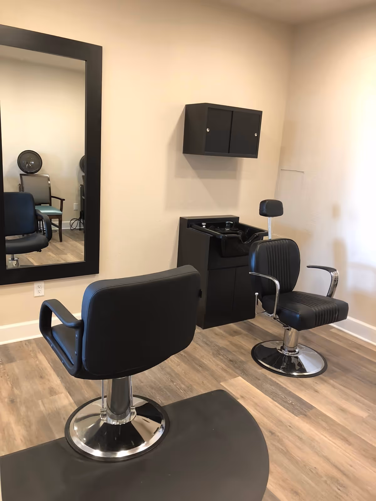 Interior of a salon area with two black salon chairs, a black hair washing station, a large wall mirror, and a black wall-mounted cabinet. The floor has wood-like vinyl flooring and the walls are painted beige.