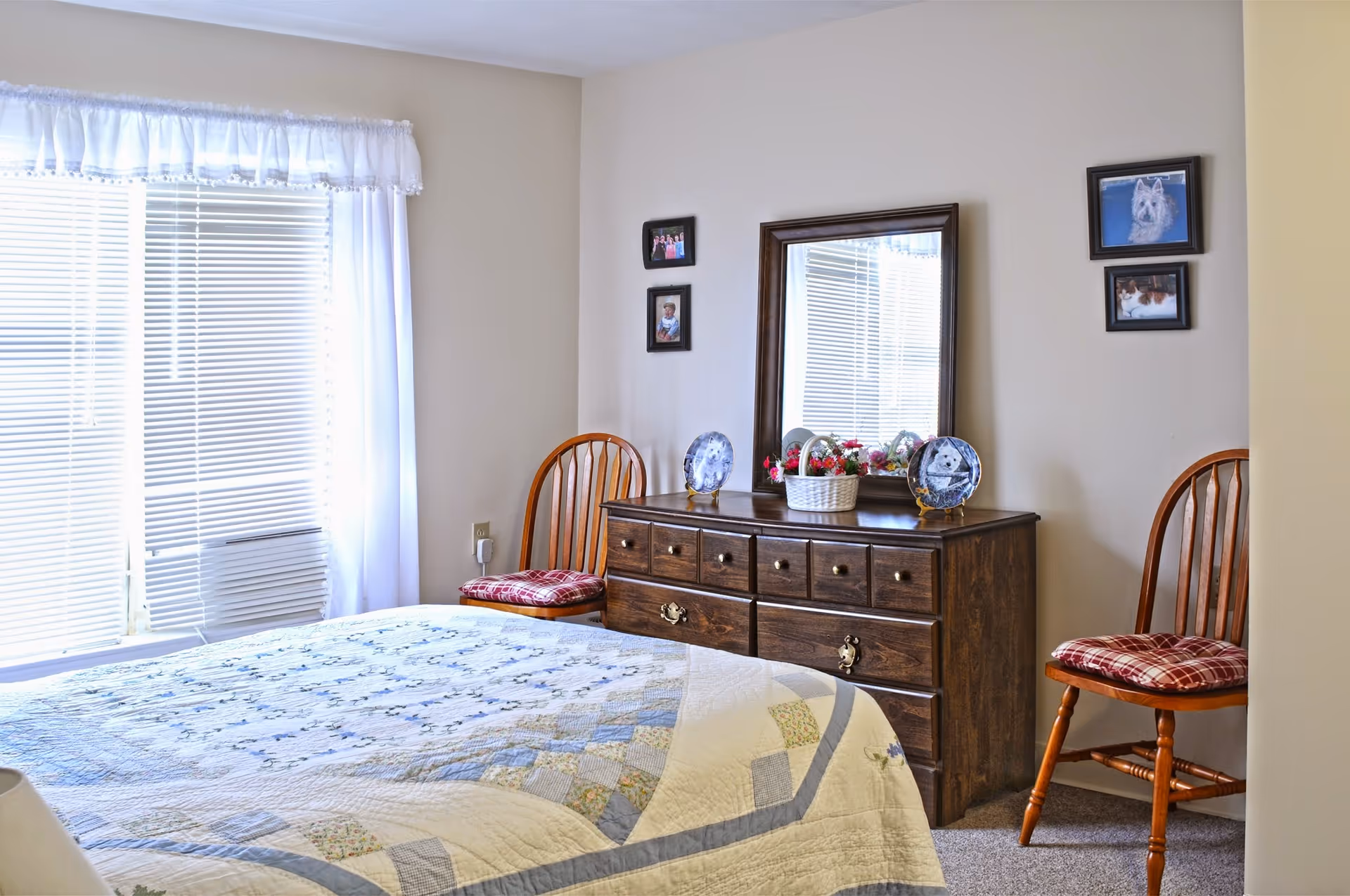 A cozy bedroom with a bed covered in a patterned quilt, two wooden chairs with red plaid cushions, a wooden dresser with a mirror, decorative plates, and a basket of flowers. The room has beige walls and a window with white blinds and sheer curtains.
