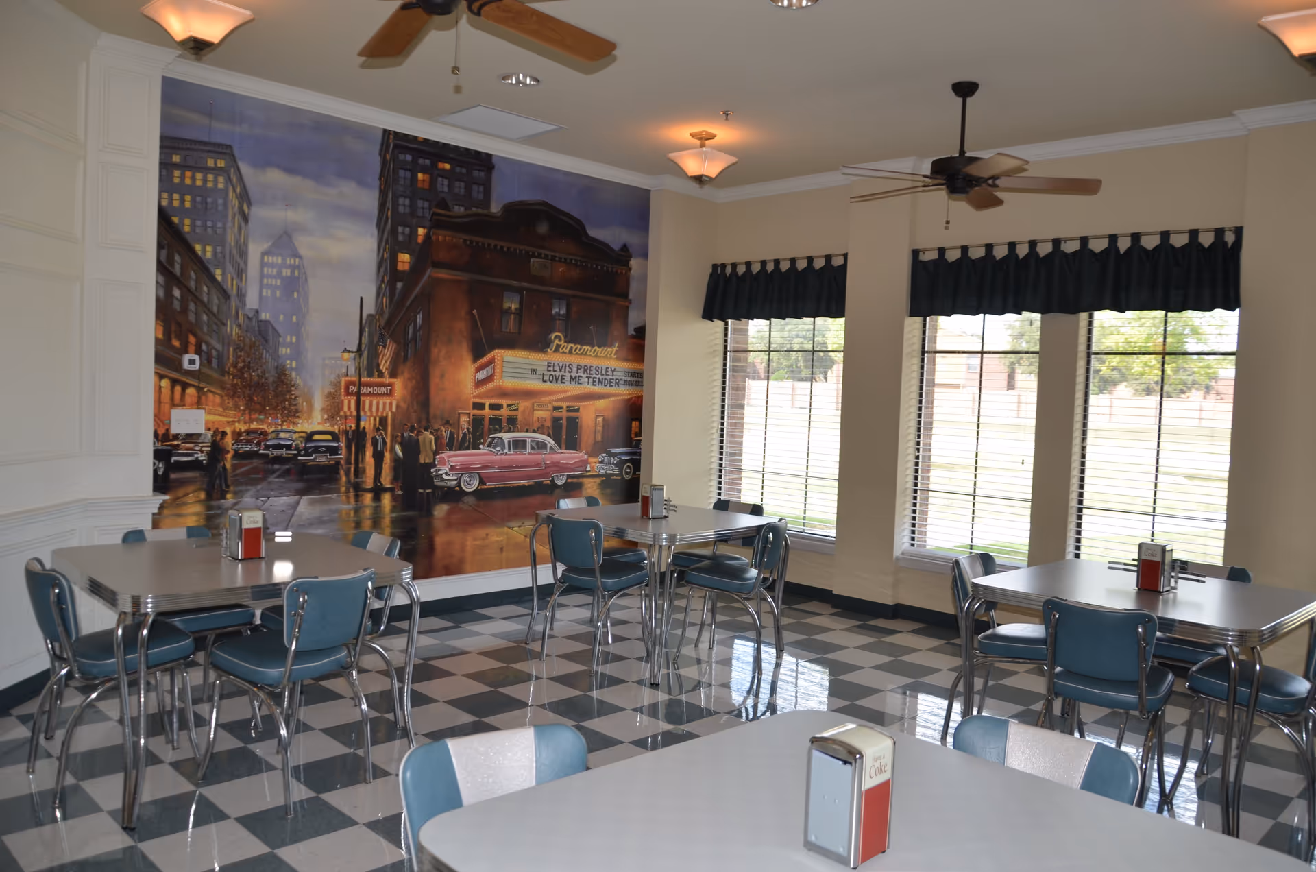 A retro-style dining room with checkered black and white floor tiles, several tables with blue and white chairs, large windows with blinds and dark valances, ceiling fans, and a mural on one wall depicting a vintage city street scene with a Paramount theater marquee showing Elvis Presley in Love Me Tender.
