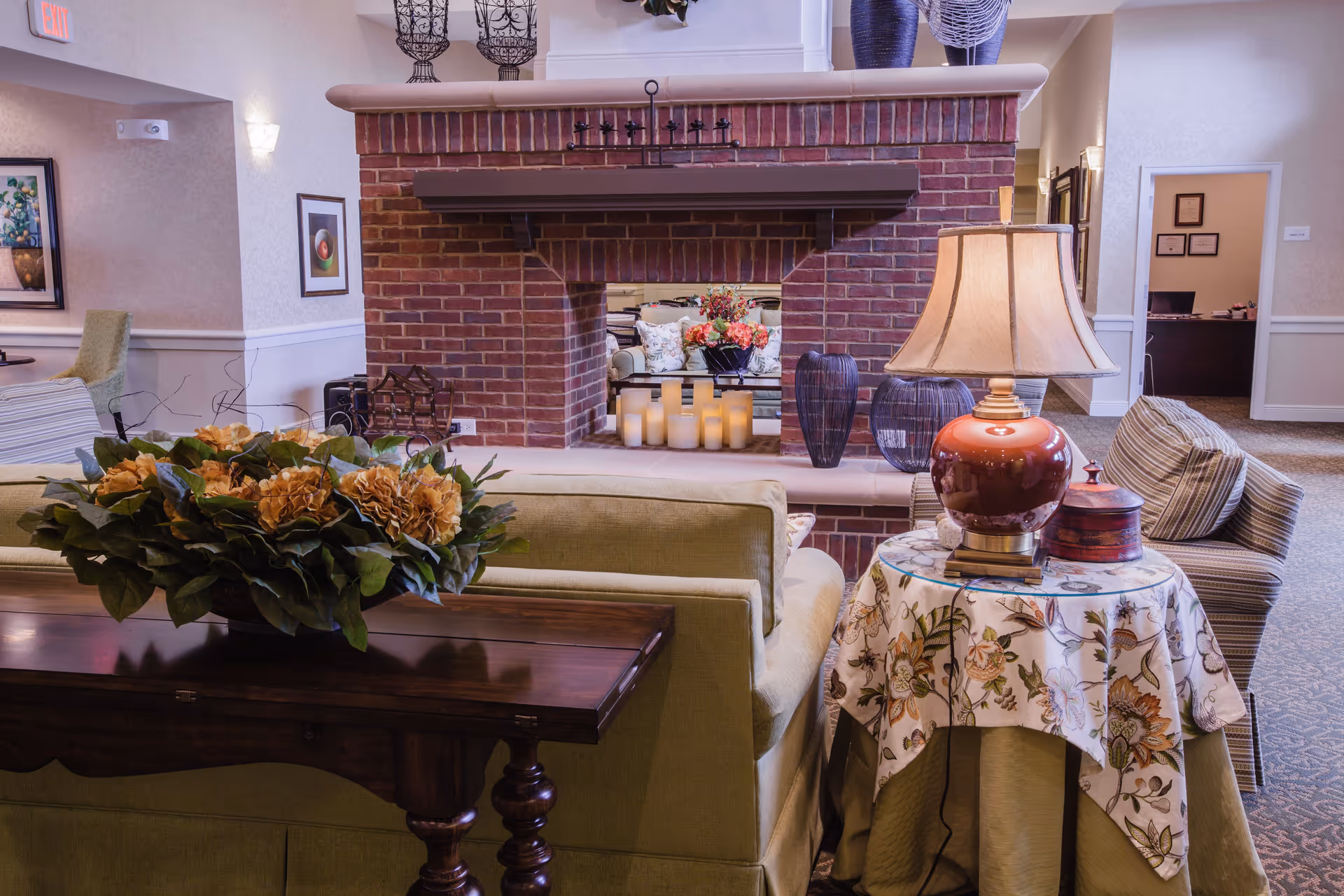 Cozy living room area in an assisted living facility featuring a brick double-sided fireplace with candles inside, a green sofa with a wooden table behind it holding a floral arrangement, a round side table with a floral tablecloth, a red lamp, and striped armchairs. The room has soft lighting, framed artwork on the walls, and an open doorway leading to an office area.