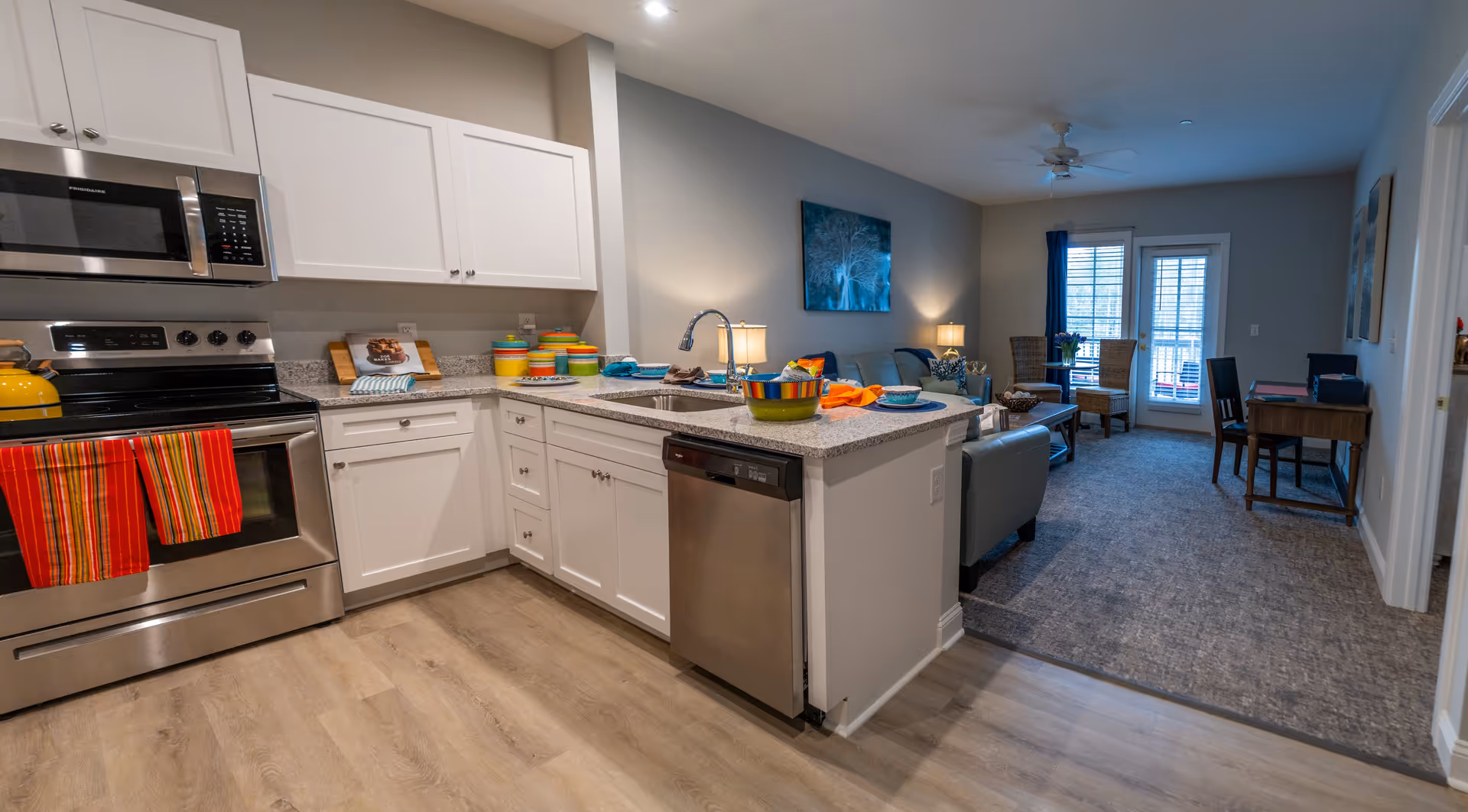 Modern kitchen with white cabinets, stainless steel appliances including a microwave, stove, and dishwasher. The kitchen island has a sink and colorful bowls and towels. The kitchen opens into a living area with a sofa, coffee table, chairs, a desk, and a door leading to a balcony or patio.