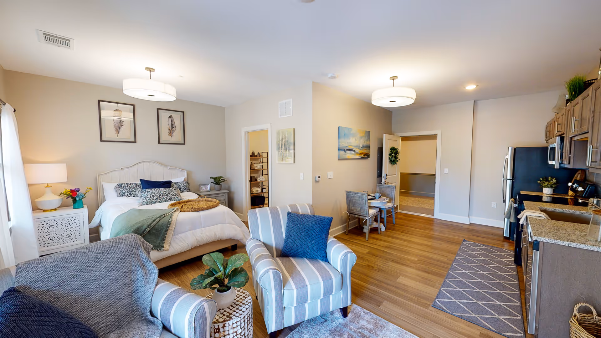 A bright and spacious studio apartment featuring a cozy bed with white bedding and decorative pillows, two framed feather artworks above the bed, a bedside table with a lamp and flowers, a striped armchair with a blue cushion, a small round table with a plant, a dining area with a small table and two chairs, and a modern kitchen with wooden cabinets, granite countertops, and stainless steel appliances. The room has wood flooring and neutral-colored walls with additional artwork.