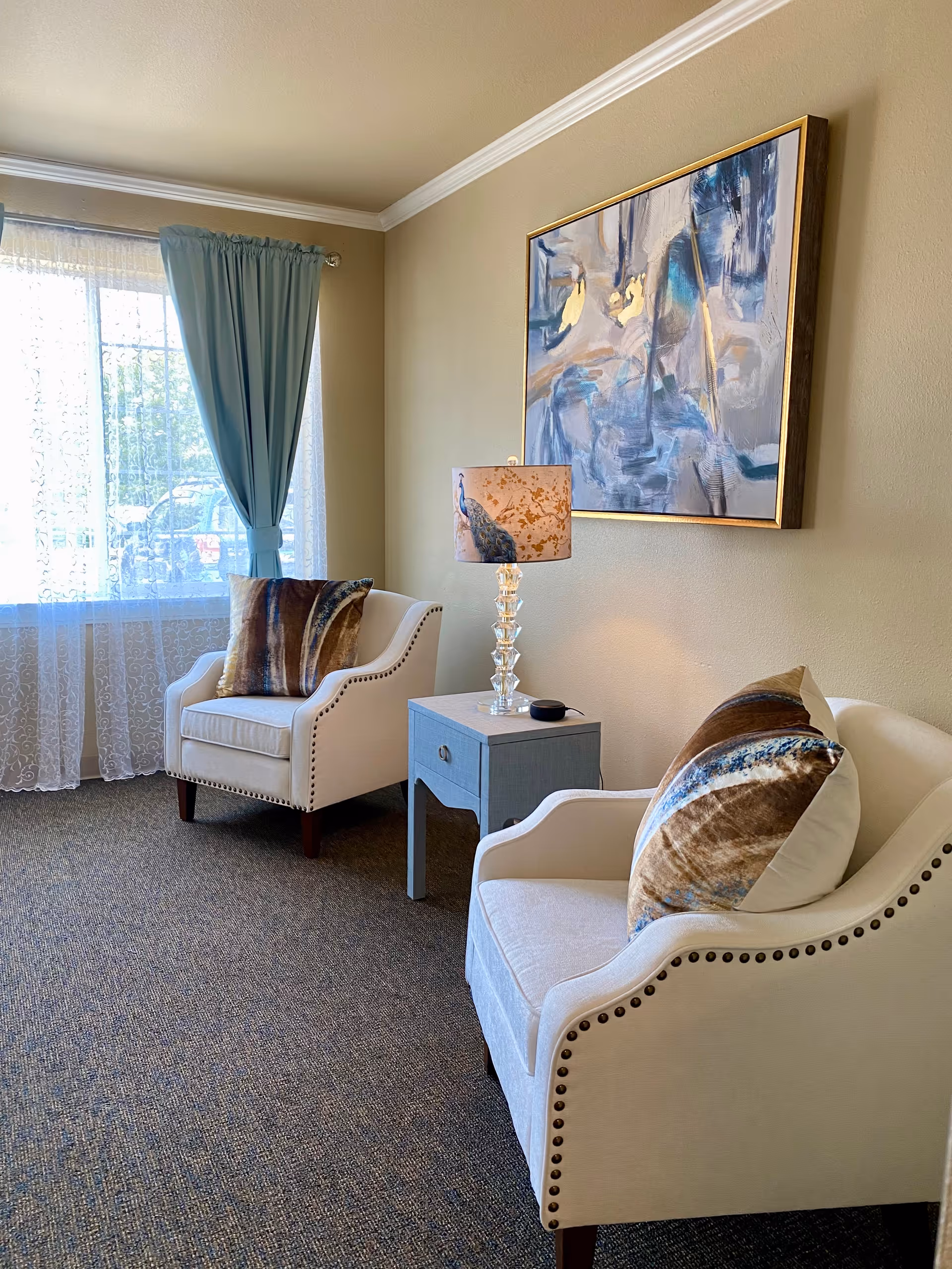 A cozy sitting area with two cream-colored armchairs featuring nailhead trim and decorative pillows. Between the chairs is a small blue side table with a glass lamp that has a peacock design on the lampshade. A large abstract painting with blue and beige tones hangs on the wall above the table. A window with sheer white curtains and blue drapes allows natural light into the room.