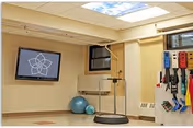 Rehabilitation exercise room with a wall-mounted TV, balance platform, exercise balls, and assorted resistance bands.