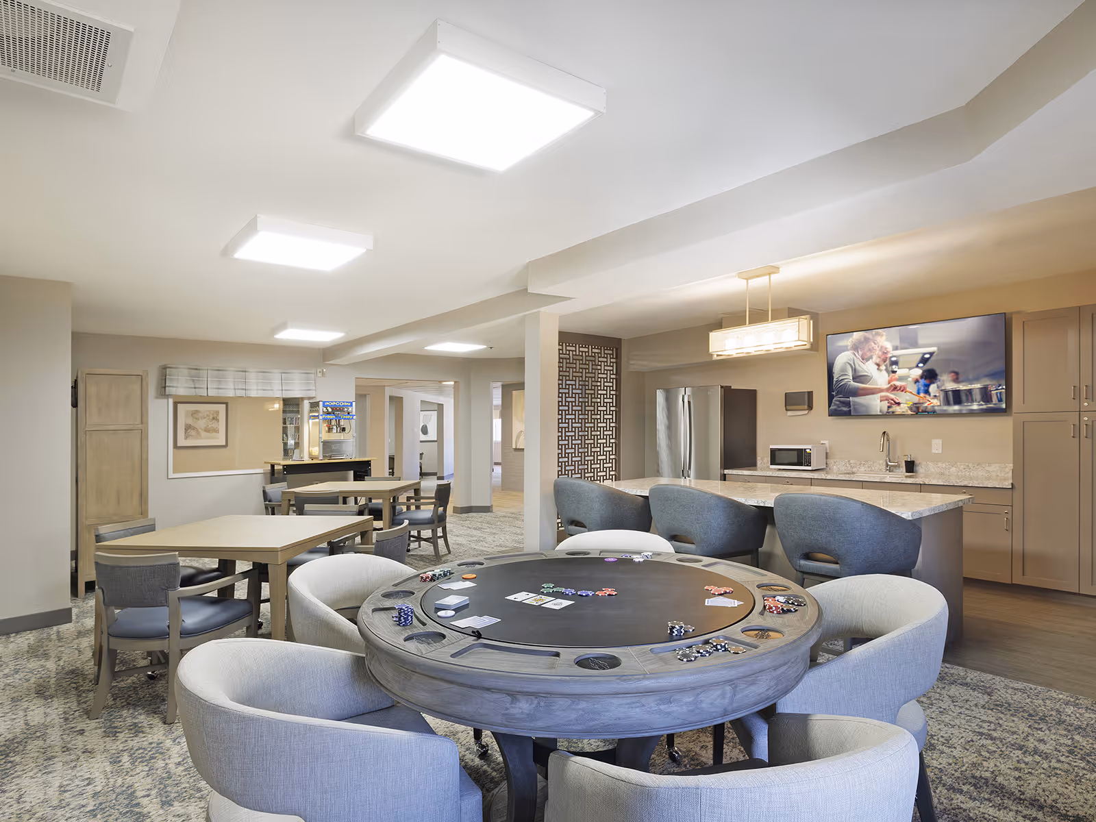 A spacious common area in a senior living facility featuring a round poker table with chips and cards, surrounded by cushioned chairs. In the background, there are additional tables and chairs, a popcorn machine, a kitchen area with a refrigerator, microwave, and a countertop with bar stools. A large TV mounted on the wall displays an image of people cooking.