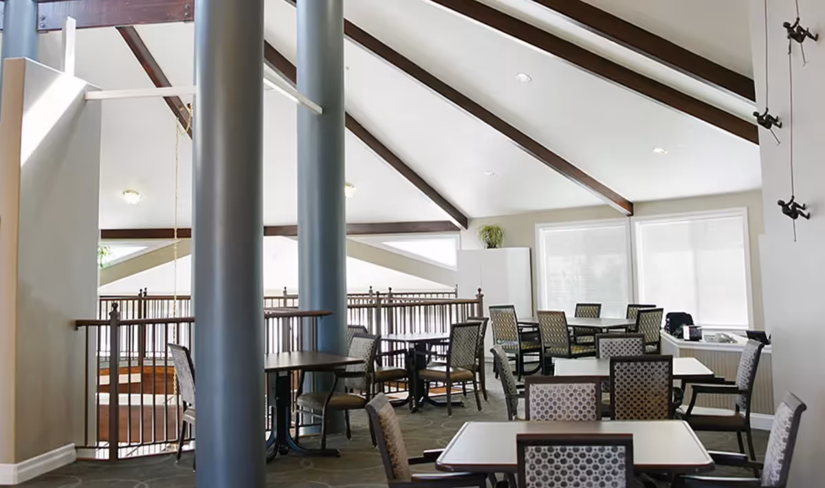 Interior of a dining area with multiple tables and chairs arranged neatly. The room features a high ceiling with exposed wooden beams and large windows letting in natural light. There are two large gray columns in the middle of the space and a railing along one side.