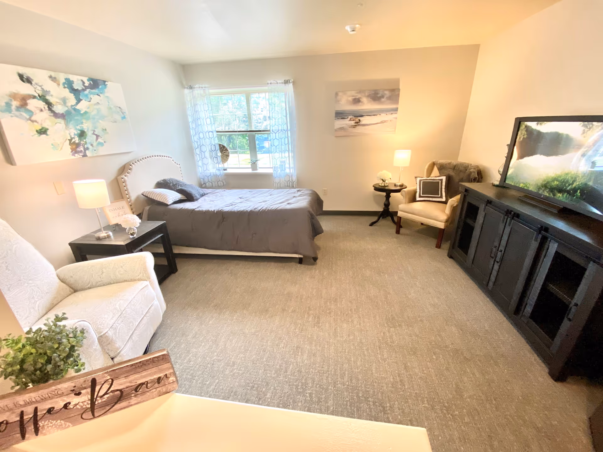A cozy bedroom in a senior living facility featuring a single bed with gray bedding, two armchairs, a black TV stand with a flat-screen TV, a window with patterned curtains, two side tables with lamps, and wall art depicting abstract and beach scenes.