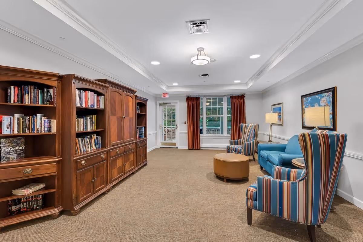 A cozy living room area with a large wooden bookshelf filled with books on the left side, and a seating area on the right featuring a blue sofa, two striped armchairs, a round ottoman, and a side table with a lamp. The room has beige carpet, white walls, a window with red curtains, and ceiling lights.
