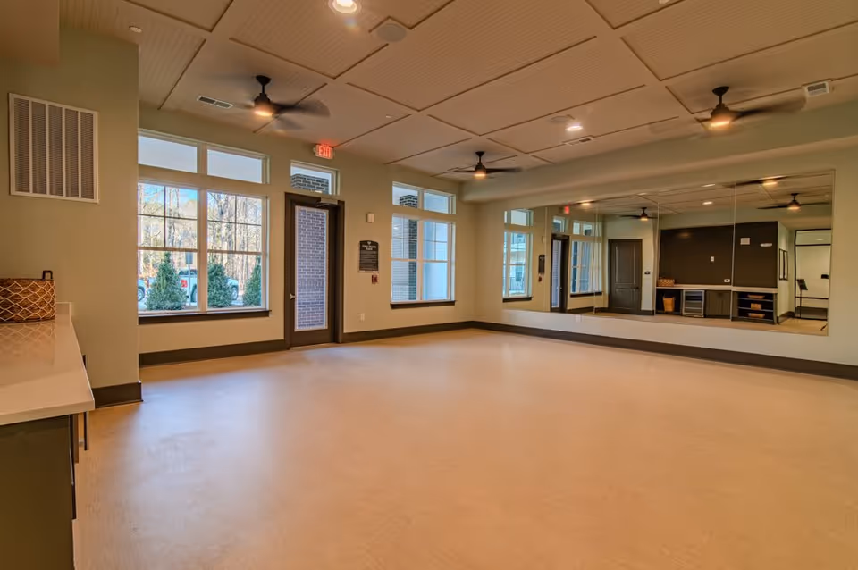 A spacious, empty room with large windows letting in natural light, a door leading outside, ceiling fans, recessed lighting, and a large wall mirror reflecting the room. The room has light-colored flooring and walls with a countertop and cabinets on one side.