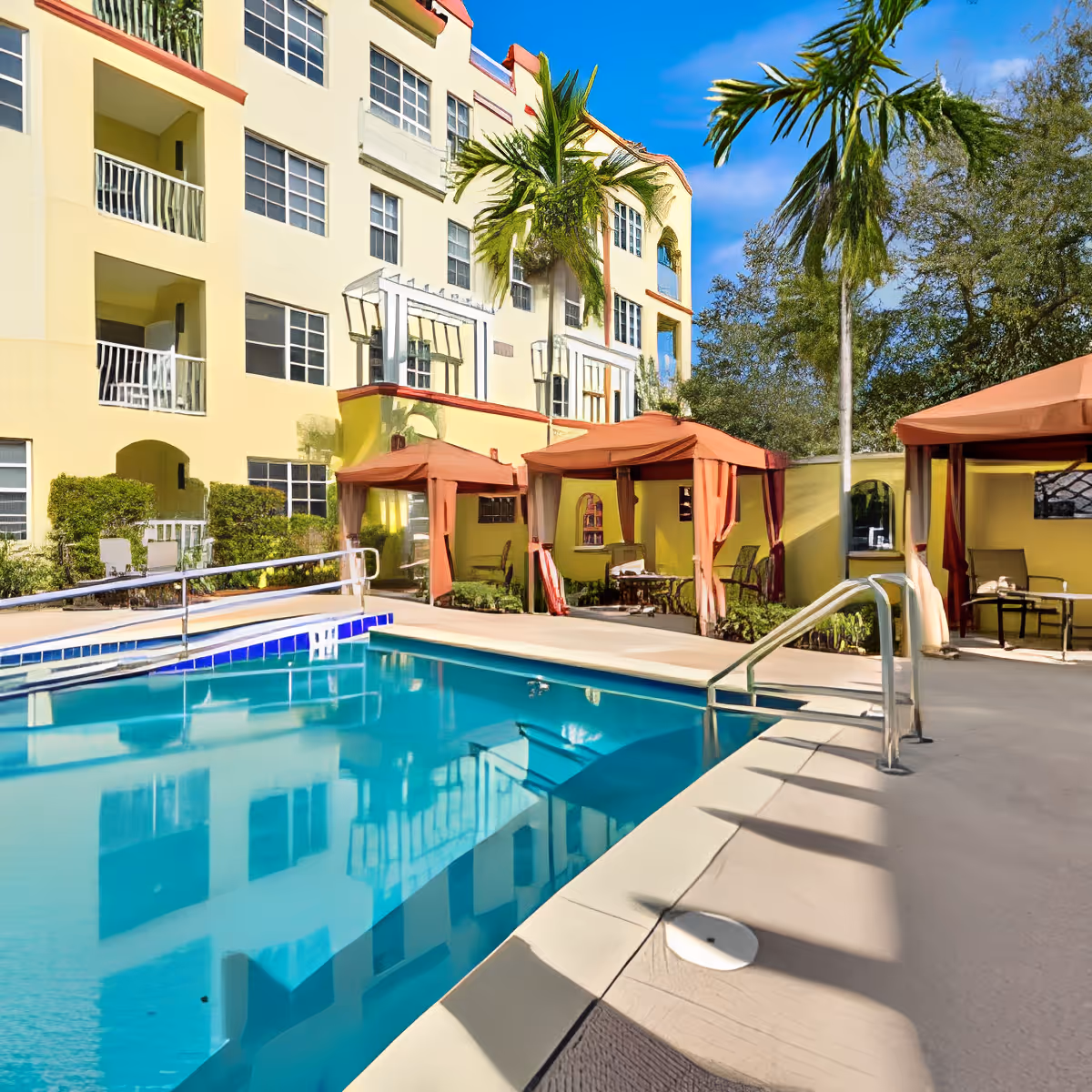 Outdoor swimming pool area at Discovery Village At Melbourne with clear blue water, surrounded by a beige concrete deck. There are three orange cabanas with seating and tables along one side of the pool. The background features a multi-story yellow building with balconies and windows, palm trees, and a bright blue sky.