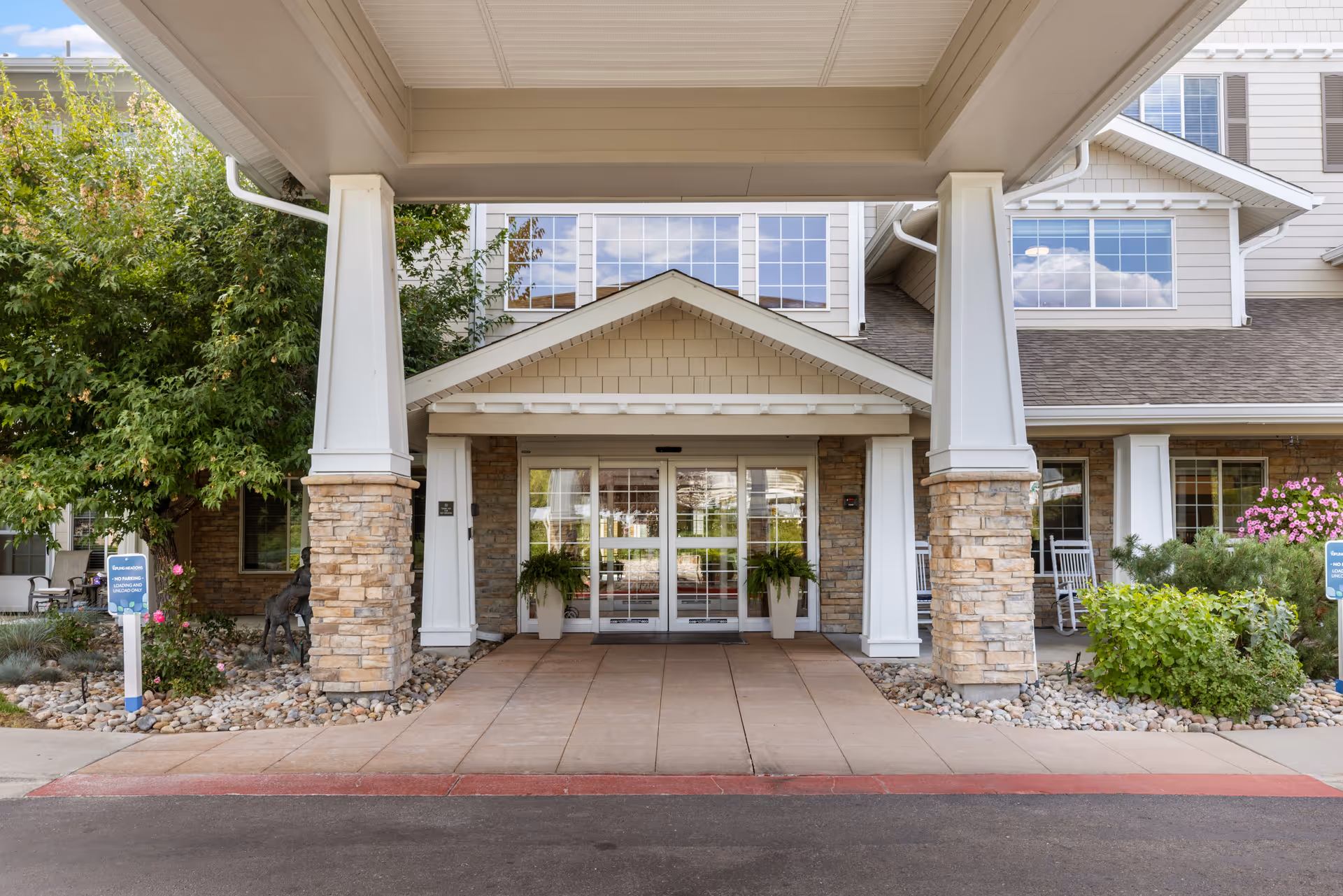 Covered main entrance of a senior living building with stone columns, a porte-cochère, glass automatic doors, potted plants, and a driveway.