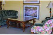 A cozy living room area with two green upholstered sofas, one with a floral pattern, and a wooden coffee table in the center. Two framed pictures hang on the light yellow wall behind the sofas, and there are two table lamps on either side of the room.