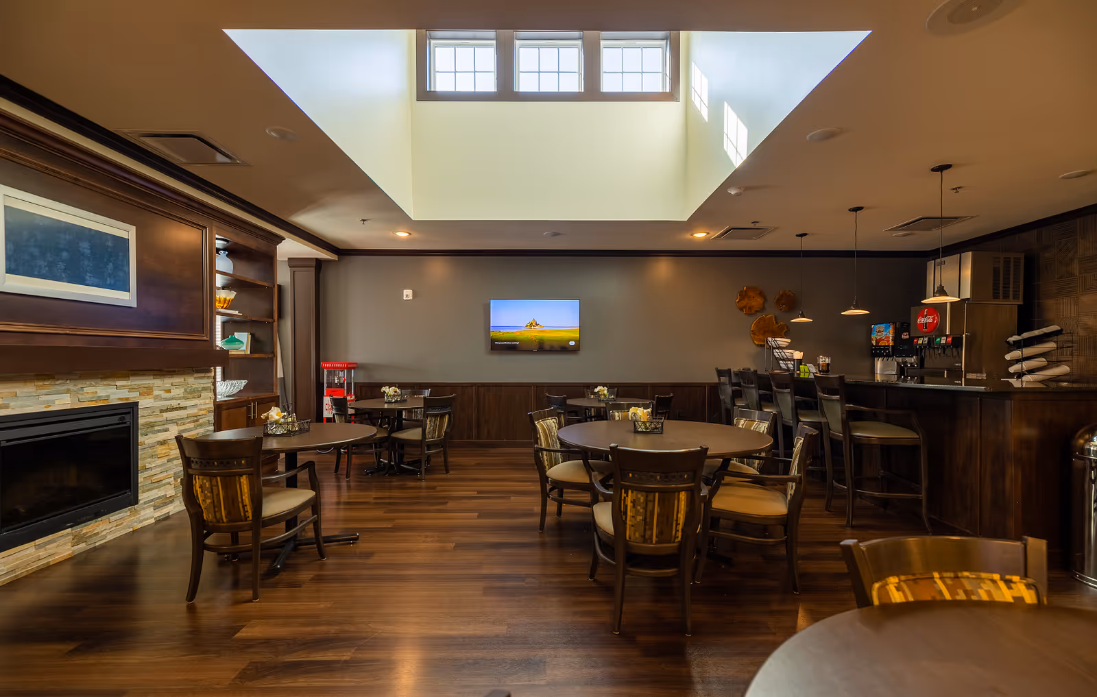 Well-lit communal dining and lounge area with round tables, chairs, a bar, fireplace, and a large skylight.