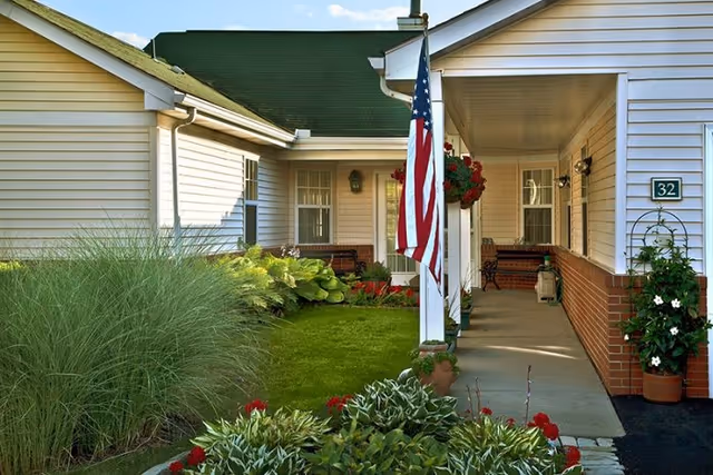 Front exterior of a single-story retirement community with flag, porch seating, and plants