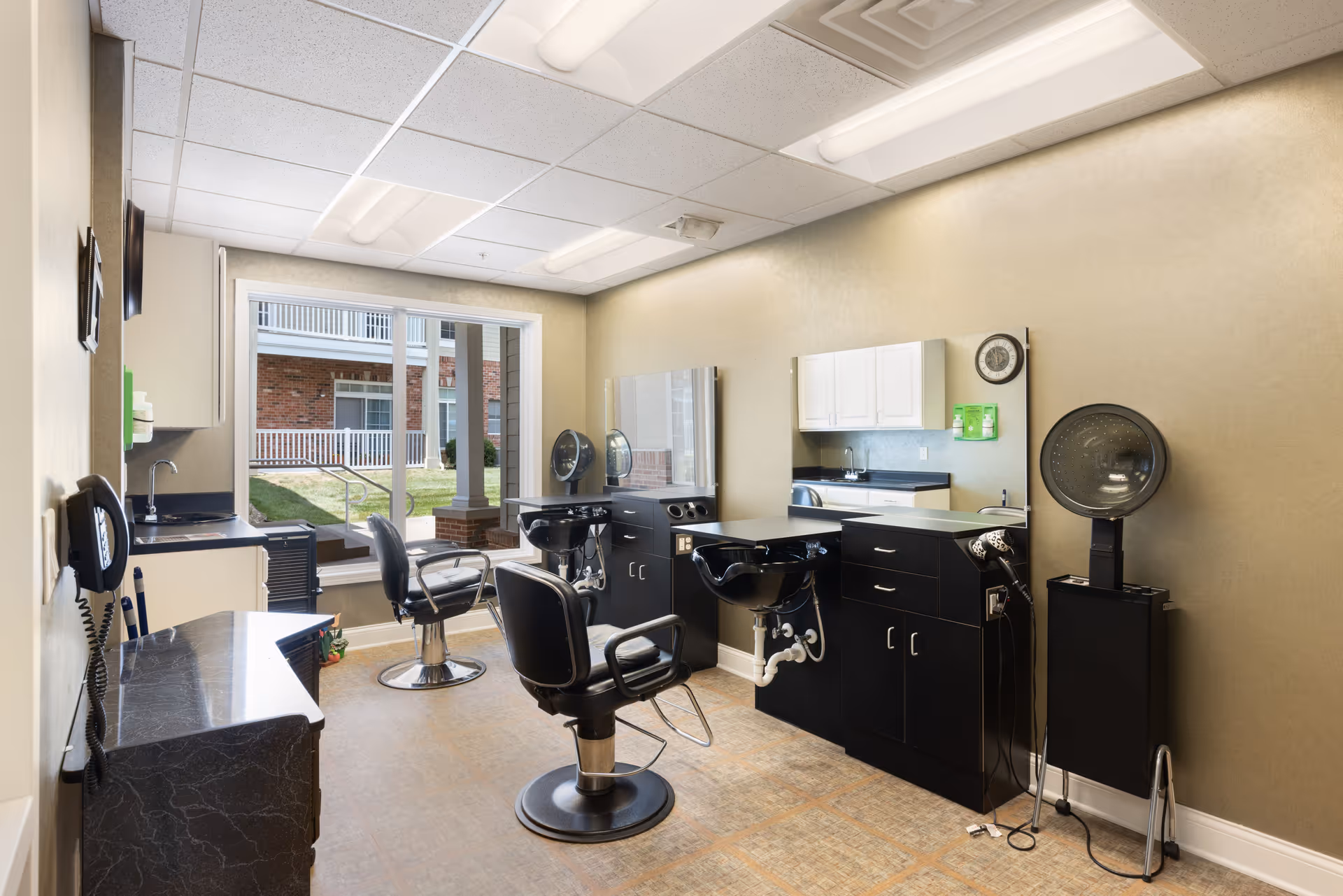 Interior view of a salon room in a senior living facility with two black salon chairs, two black wash basins, large mirrors on the wall, a hair dryer, and a window showing an outdoor area with grass and a brick building.