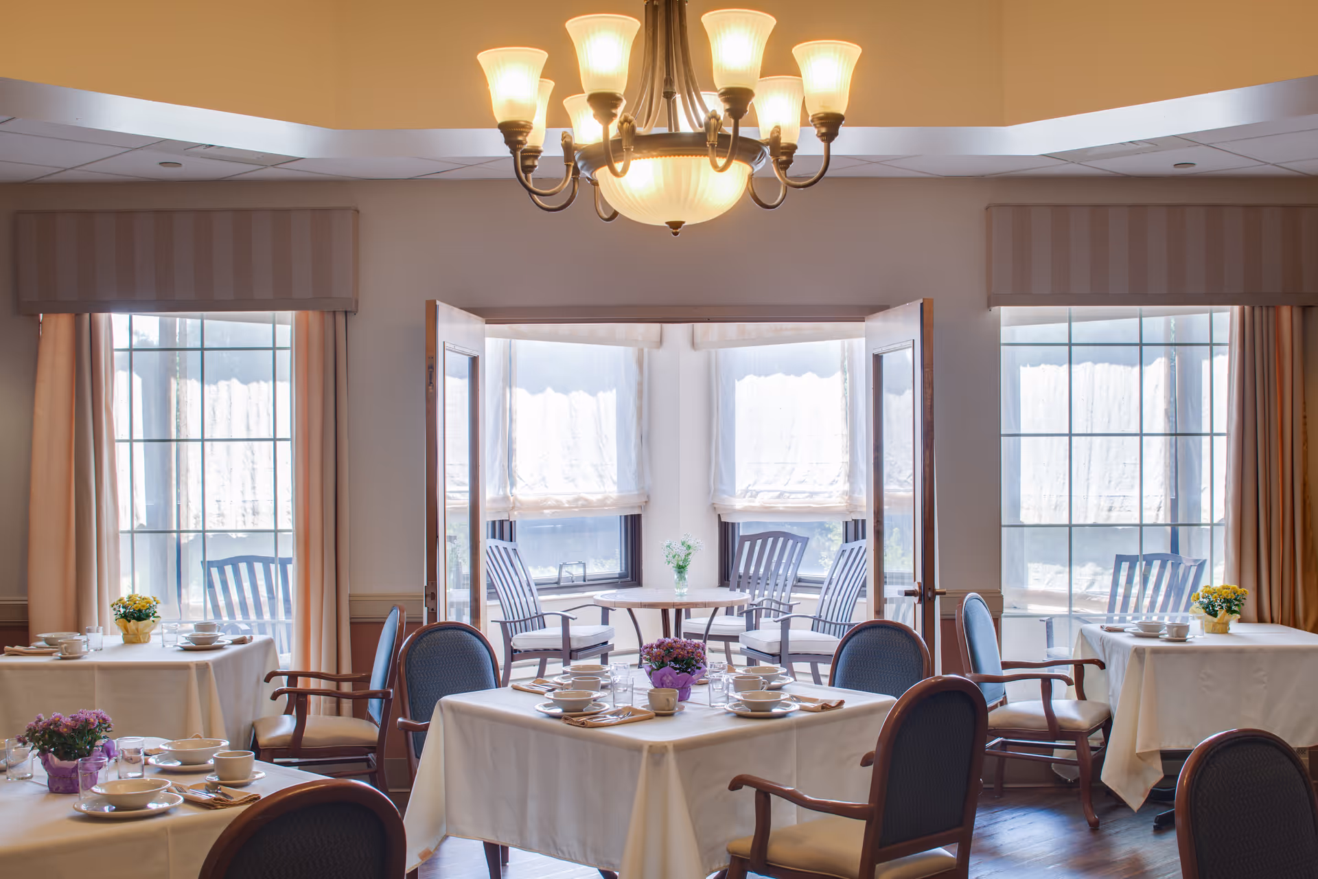 A bright dining room with several tables covered in white tablecloths, each set with plates, cups, and utensils. The room has large windows with beige curtains and a chandelier hanging from the ceiling. There are wooden chairs with cushions around the tables and a small table with chairs near the windows.