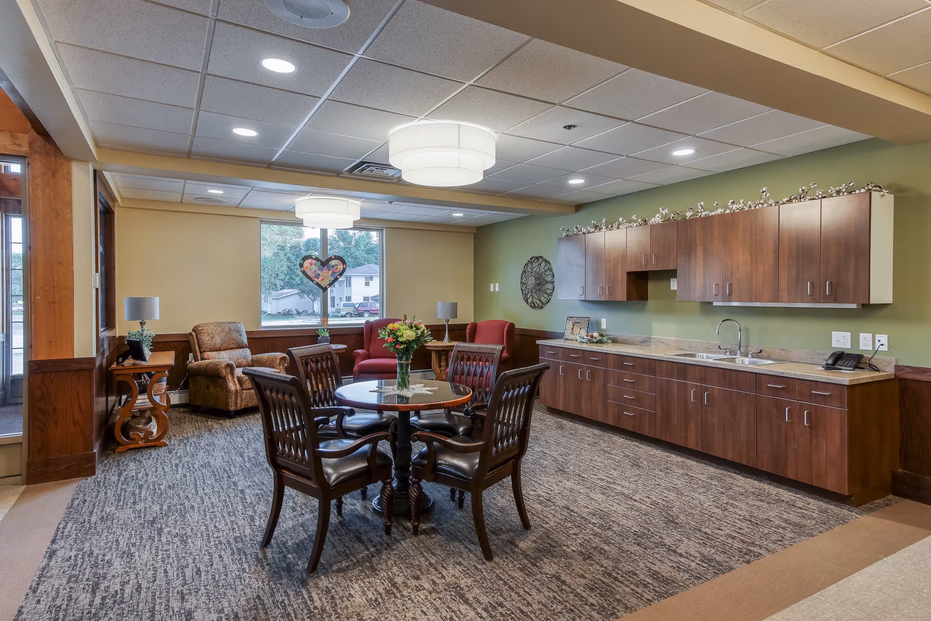 A cozy senior living common area with a round table and four wooden chairs in the center. There are two armchairs and a side table with a lamp near a large window. The room features a long countertop with a sink and wooden cabinets above and below. The walls are painted green and beige, and the ceiling has recessed lighting and two round ceiling lights.