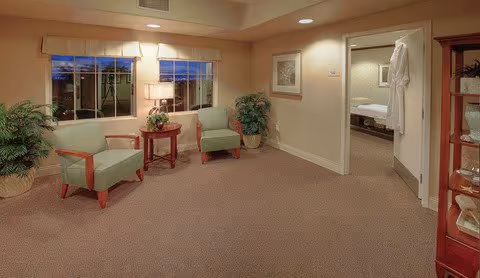 A cozy interior room with two green armchairs and a small round wooden table between them, placed near large windows with a view outside. The room has beige walls and carpet, with potted plants and framed artwork on the walls. An open doorway leads to a bedroom with a visible bed and a white robe hanging on the door frame.