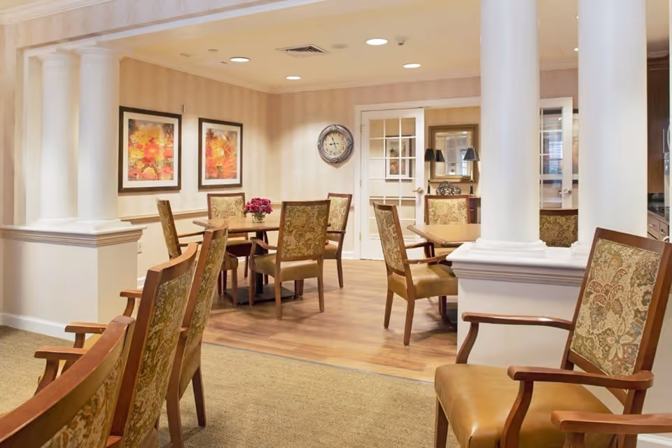 A well-lit interior room with several wooden chairs featuring patterned upholstery arranged around wooden tables. The room has beige walls with two framed floral paintings, a wall clock, white columns, and a glass-paneled door leading to another room.