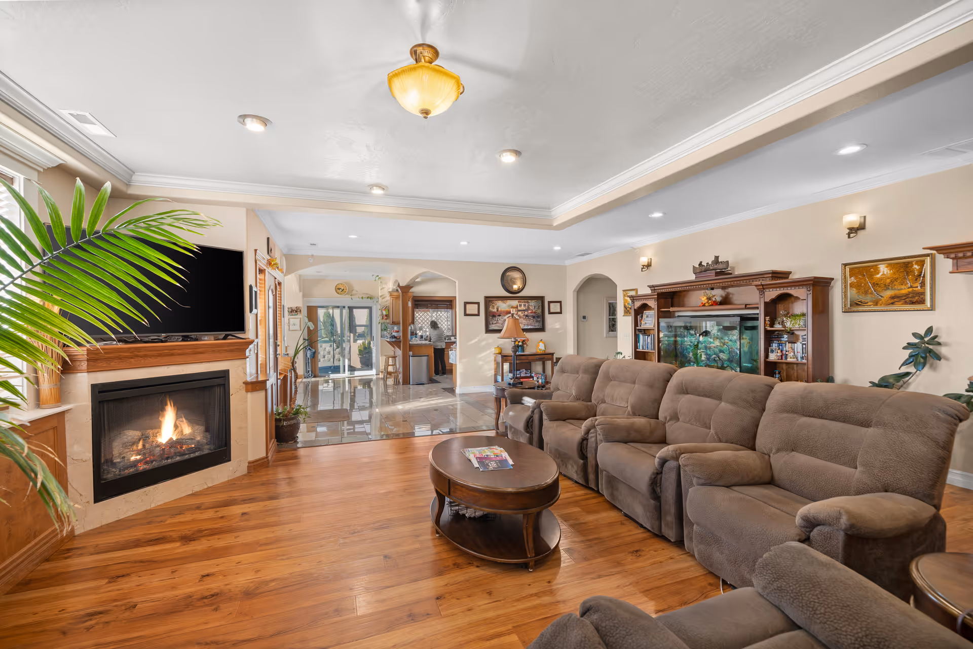 A cozy living room with a brown reclining sofa set arranged around a wooden coffee table. There is a fireplace with a flat-screen TV mounted above it on the left side. The room has wooden flooring, recessed ceiling lights, and a ceiling light fixture. In the background, there is a view into a kitchen area with tiled flooring and a large fish tank built into a wooden cabinet on the right wall. Various plants and framed pictures decorate the space.