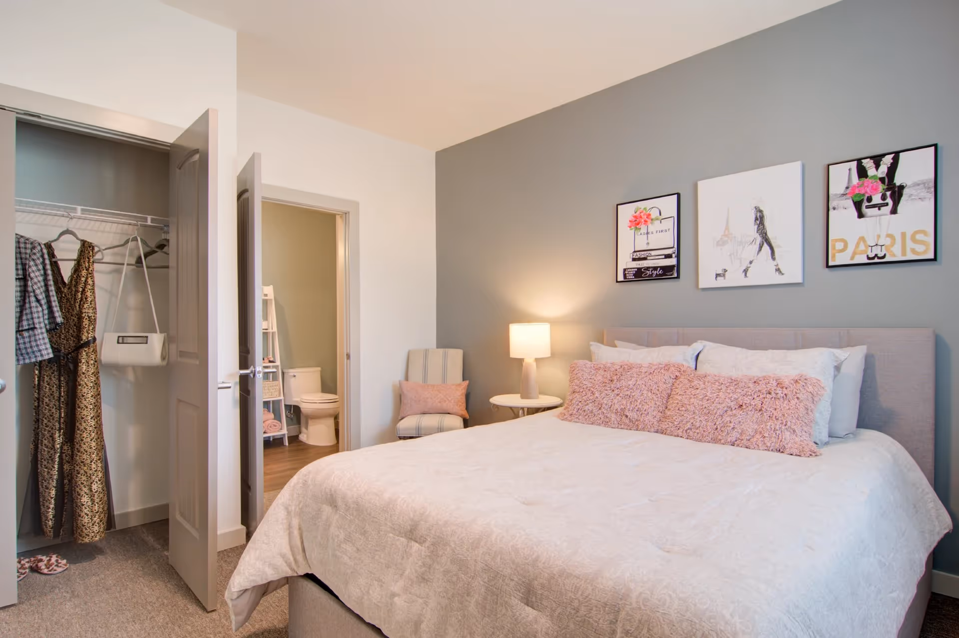 A cozy bedroom with a large bed featuring white bedding and pink fluffy pillows. On the wall above the bed are three framed art pieces with fashion and Paris themes. To the right of the bed is a small round table with a lamp and a striped chair with a pink pillow. On the left side, there is an open closet with clothes hanging and an open door leading to a bathroom with a visible toilet and shelving unit.
