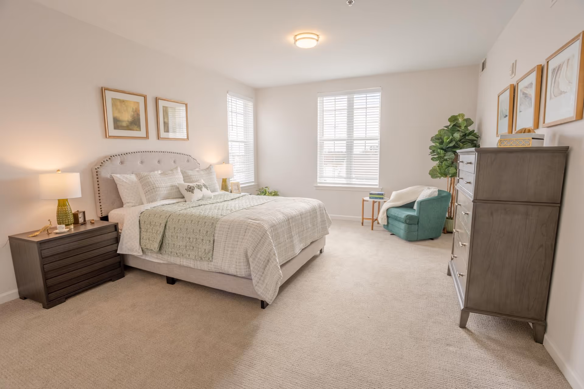 A bright and spacious bedroom with a large bed featuring a tufted headboard, light-colored bedding, and multiple pillows. There are two windows with white blinds letting in natural light. On the left side of the bed is a dark wooden nightstand with a lamp and small decorative items. On the right side of the room is a green armchair with a white throw blanket, a small wooden side table with books, a tall dark wooden dresser, and a large potted plant. The walls are light-colored with framed artwork above the bed and dresser. The floor is carpeted in a neutral tone.