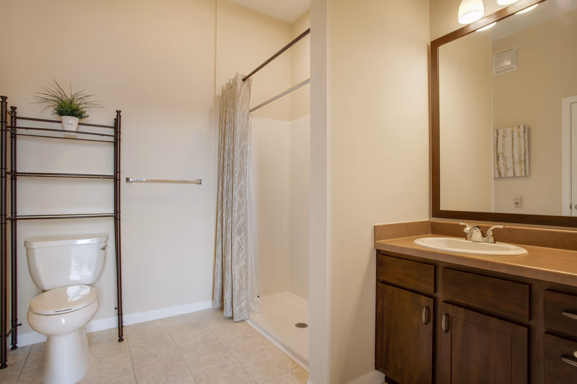 A clean bathroom featuring a white toilet with a black metal shelving unit above it holding a small potted plant. Next to the toilet is a walk-in shower with a beige patterned curtain. On the right side, there is a wooden vanity with a brown countertop, a white sink, and a large mirror above it. The walls are painted light beige and the floor is tiled.