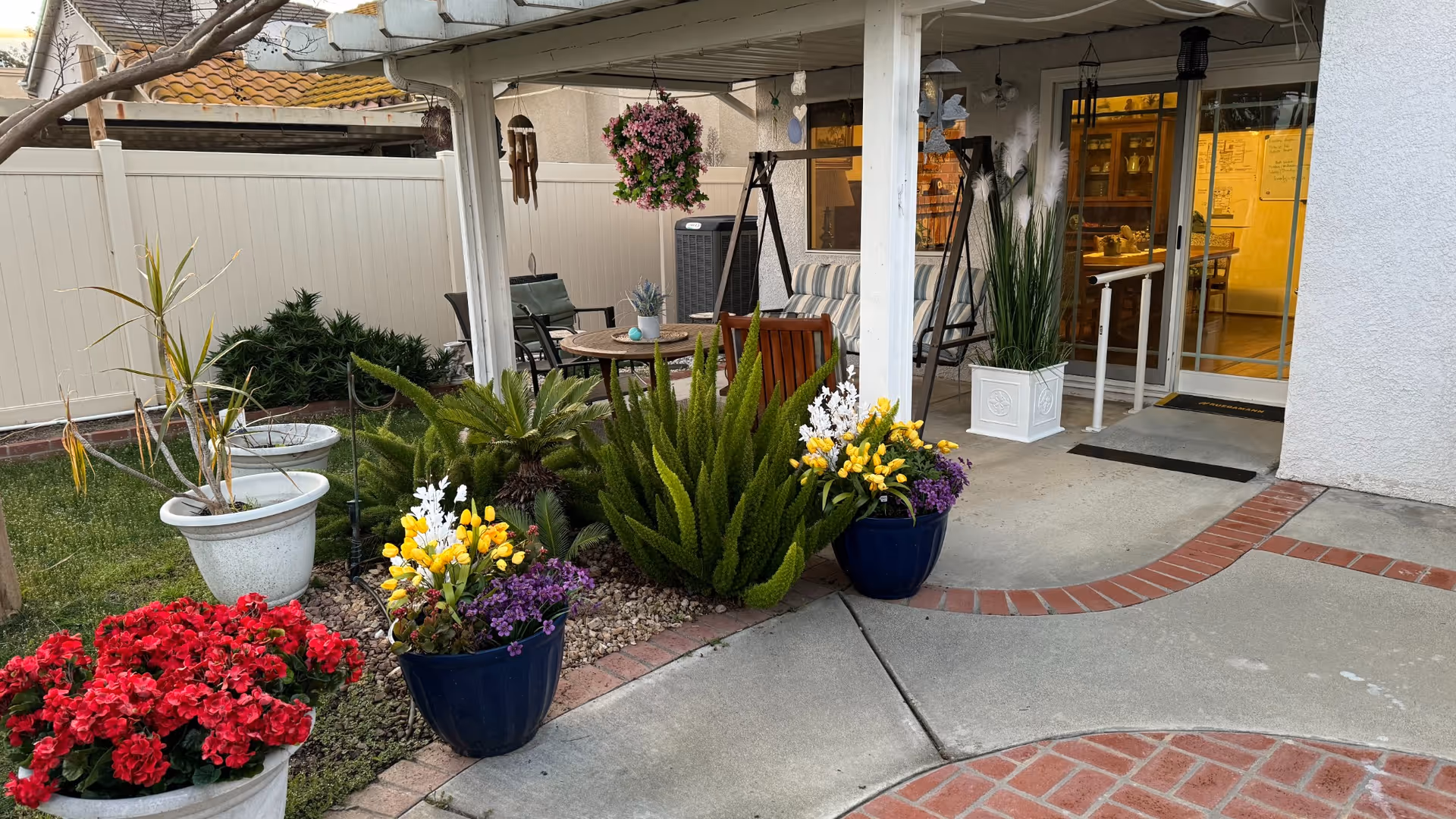Outdoor patio area with a covered seating space featuring a swing bench and chairs around a small table. The patio is surrounded by various potted plants and flowers, including red, yellow, white, and purple blooms. There is a white fence in the background and a sliding glass door leading inside the building.