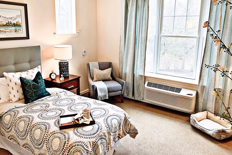 A cozy bedroom with a bed featuring a patterned bedspread and multiple pillows, a wooden nightstand with a lamp and clock, a gray armchair with a pillow and throw, large windows with light blue curtains, an air conditioning unit below the window, and a small pet bed on the carpeted floor.