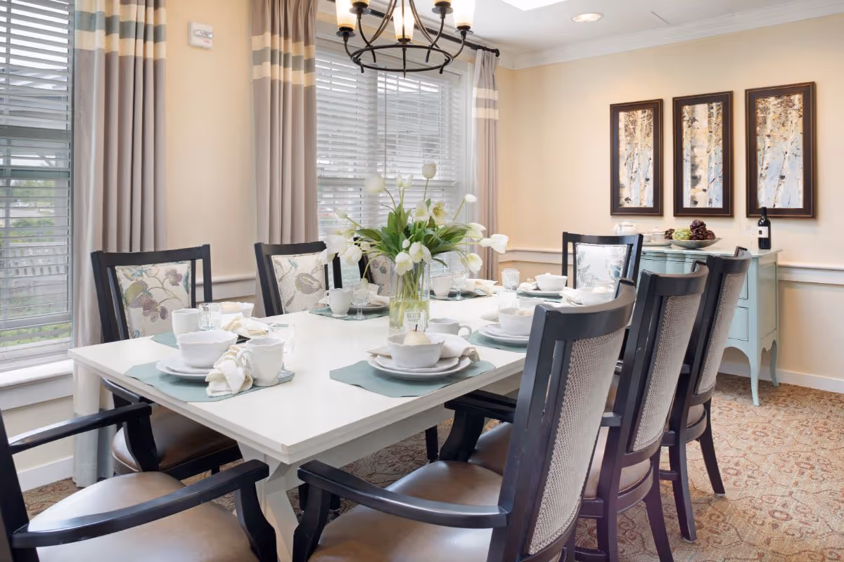 Bright formal dining room with a white table set for six, floral centerpiece, and framed artwork on the wall.