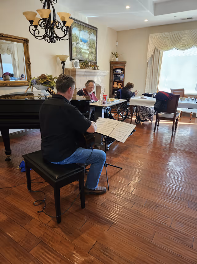 A man playing guitar while seated on a bench in a room with wooden floors. Two elderly individuals are seated at tables, one in a wheelchair, engaged in activities. The room features a grand piano, a chandelier, a fireplace with a painting above it, and large windows with curtains allowing natural light to enter.