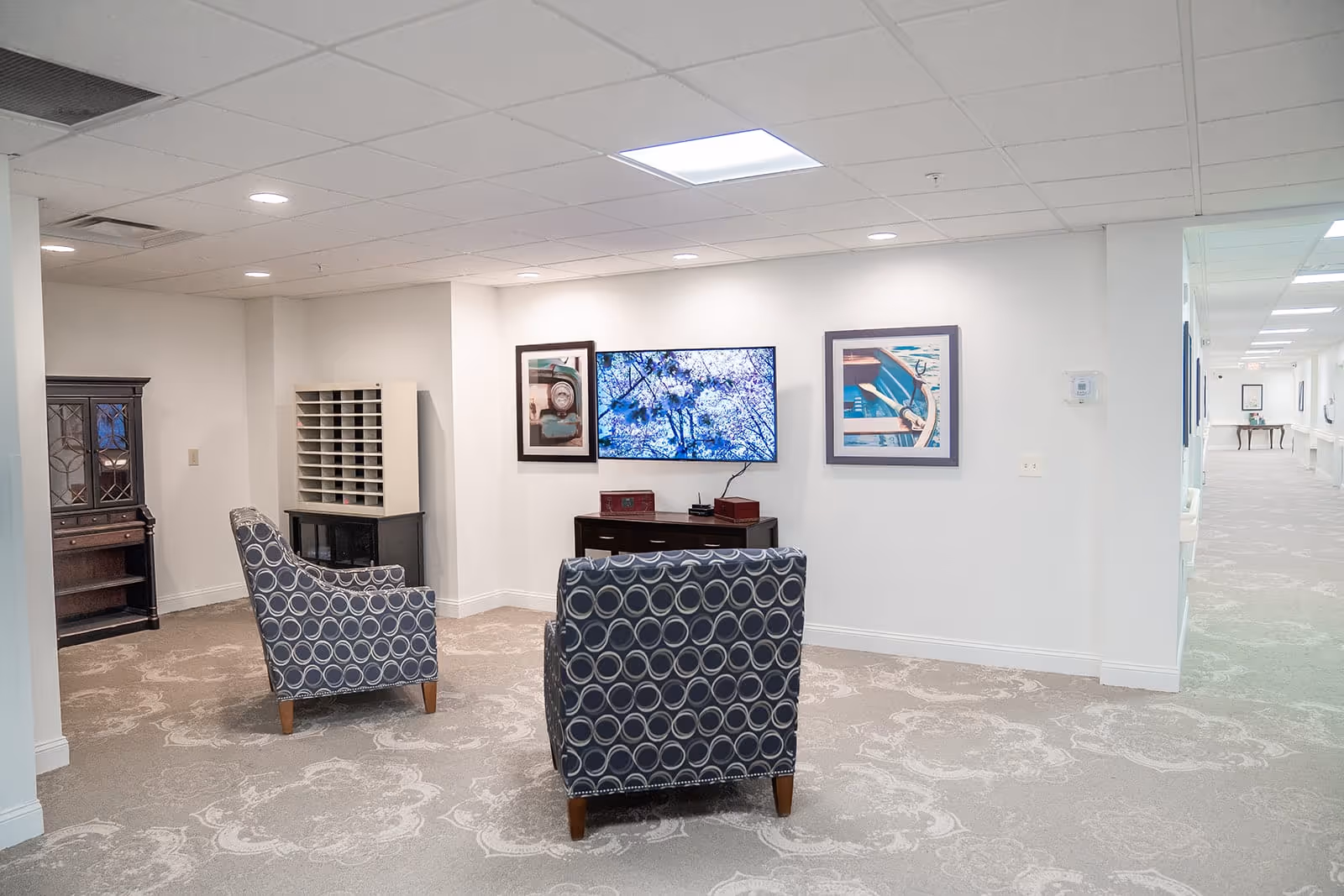 A cozy interior common area with two patterned armchairs facing a wall-mounted flat screen TV. The room has light-colored walls and carpet with a floral pattern. There are framed pictures on the wall beside the TV, and a wooden cabinet with multiple compartments is visible in the corner. A hallway extends to the right with additional framed pictures on the walls.