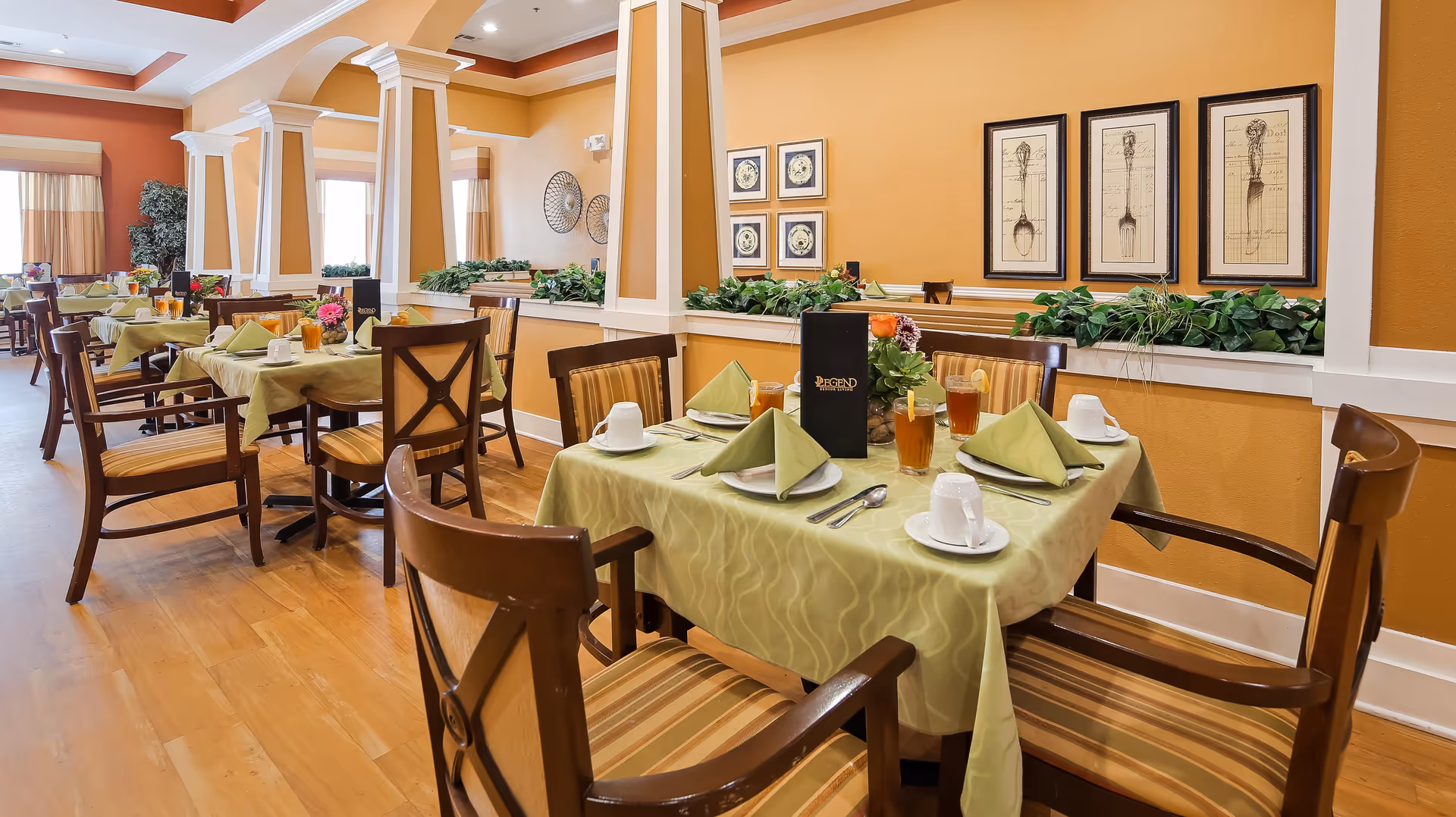A dining room with multiple tables set for meals, each covered with a light green tablecloth and neatly arranged with plates, cups, utensils, and folded napkins. The room features wooden chairs with striped cushions, warm orange walls adorned with framed artwork, and decorative plants along a ledge. Large windows with curtains allow natural light to brighten the space.