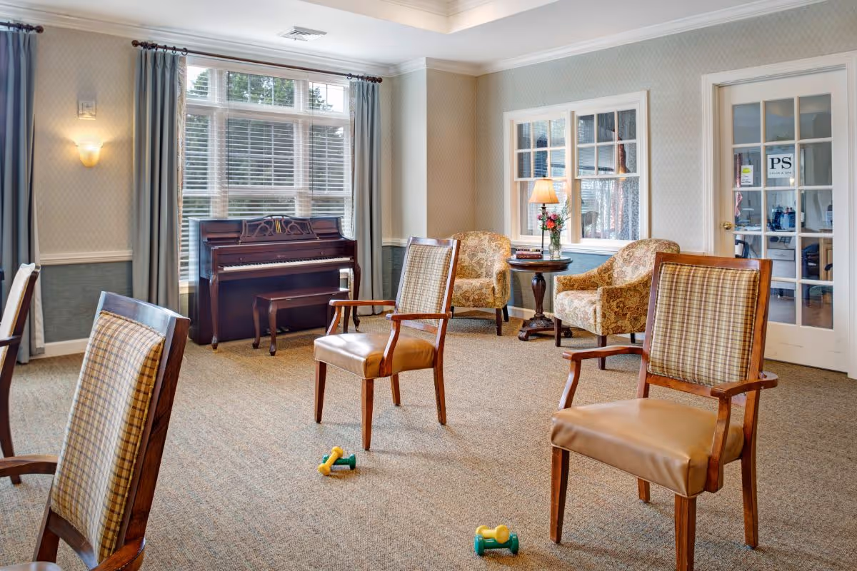 A bright senior living common room with armchairs, a piano by the window, a side table with a lamp, and small hand weights on the carpet.