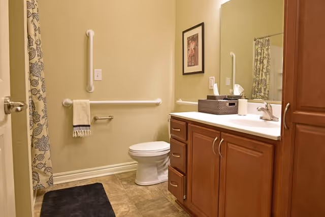 A bathroom with beige walls featuring a toilet with grab bars on the wall behind and to the side. There is a wooden vanity with a white countertop, a sink, a tissue box, and a roll of toilet paper. A framed picture hangs on the wall above the grab bar. A shower curtain with a floral pattern is partially visible on the left side, and a dark bath mat is on the tiled floor.