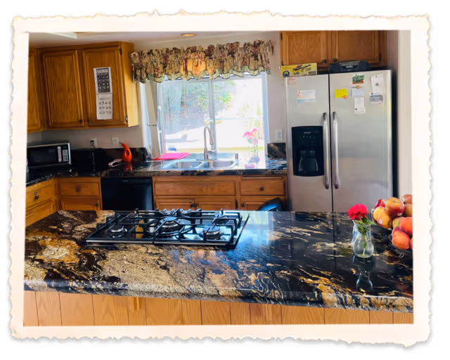 A kitchen with wooden cabinets, a black and brown marble countertop island with a built-in gas stove, a double sink under a window with floral curtains, a stainless steel refrigerator with magnets and notes, a microwave, and a small vase with a red flower and a bowl of apples on the counter.