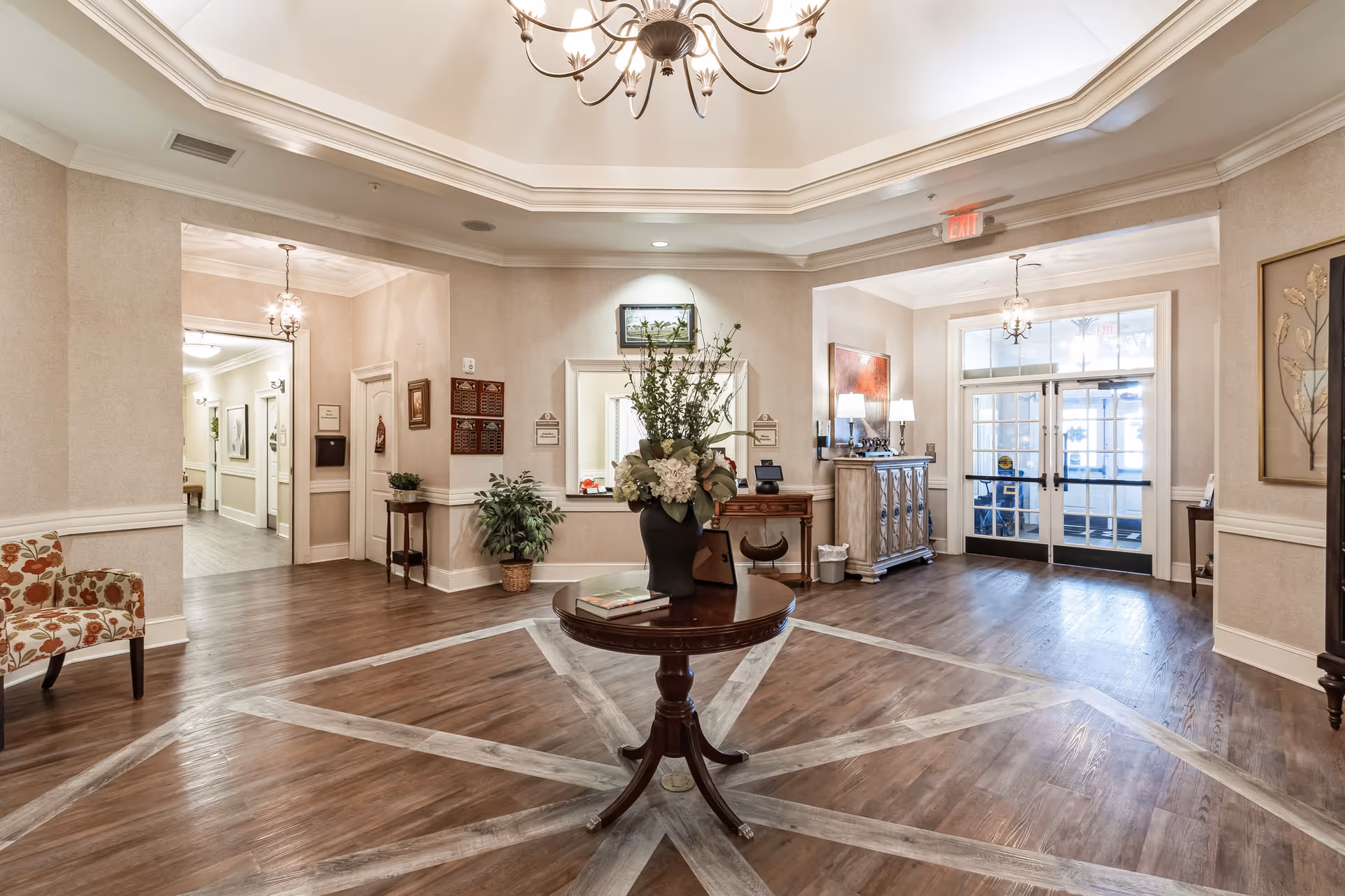 Spacious and elegant interior lobby area with a round wooden table in the center holding a large floral arrangement. The room features wood flooring with a decorative inlay, beige walls, and a chandelier hanging from a tray ceiling. There are chairs with floral upholstery, framed artwork on the walls, and double glass doors leading outside.