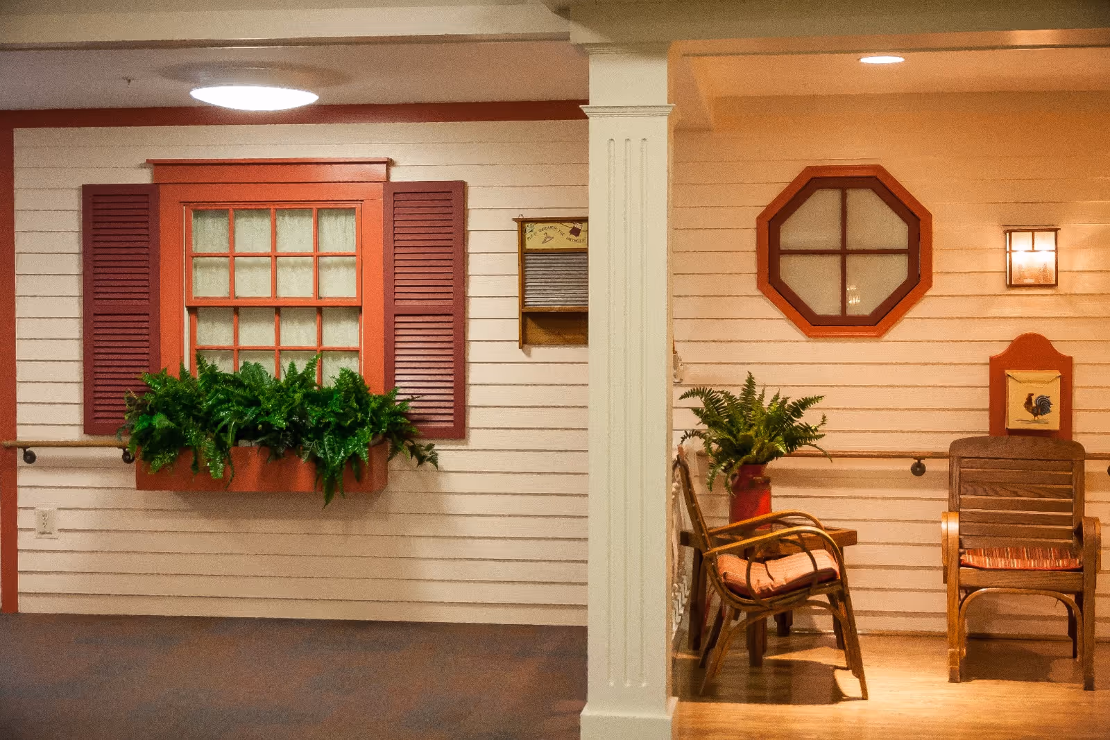 Indoor hallway area designed to resemble an outdoor porch with white siding walls, red window frames and shutters, a window box with green plants, two wooden chairs with cushions, a small table with a plant, and a wall-mounted light fixture.